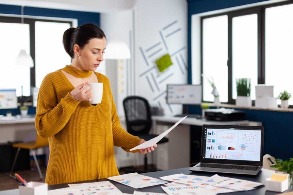Das Foto zeigt eine Geschäftsfrau in einem Büro, die konzentriert auf ein Dokument in ihrer Hand schaut. Sie trägt einen senfgelben Pullover und hält eine weiße Kaffeetasse in der Hand. Sie macht gerade eine Pause oder denkt über den Inhalt des Dokuments nach. Das Büro ist gut mit Tageslicht ausgeleuchtet und modern eingerichtet. Im Hintergrund steht ein Laptop auf einem Schreibtisch, auf dem detaillierte Geschäftsanalysen angezeigt werden, die darauf hindeuten, dass sie mit dem Facebook Business Manager arbeitet. Auf dem Tisch vor ihr liegen lose Papiere mit Diagrammen und Datenpunkten, die auf eine geschäftige Arbeitsumgebung schließen lassen. Ihr Gesichtsausdruck ist konzentriert und zeigt, dass sie mit ihrer Arbeit beschäftigt ist.