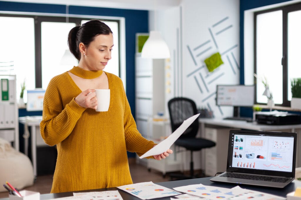 Das Foto zeigt eine Geschäftsfrau in einem Büro, in der einen Hand eine Tasse, in der anderen ein Dokument. Sie trägt einen senfgelben Pullover mit Ausschnitt, was auf eine Mischung aus professioneller und moderner Mode hindeutet. Ihr Blick ist auf das Dokument gerichtet, was darauf schließen lässt, dass sie den Inhalt überprüft oder analysiert. Hinter ihr steht ein Laptop auf einem Schreibtisch, auf dem bunte Diagramme und Grafiken zu sehen sind, die von einem Analysetool für Unternehmen wie dem Facebook Business Manager stammen könnten. Die Umgebung ist hell erleuchtet, mit einer sauberen, organisierten Büroumgebung, zu der auch ein Whiteboard mit Designelementen und Haftnotizen gehört, vielleicht für Brainstorming oder Projektplanung. Die Szene fängt einen Moment professioneller Kontemplation und Entscheidungsfindung ein.