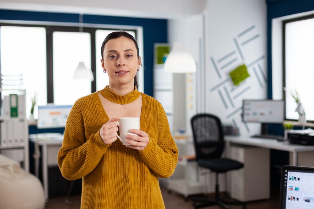 Das Bild zeigt eine Geschäftsfrau in einem Büro, die mit beiden Händen eine weiße Kaffeetasse hält. Sie wirkt entspannt und blickt mit einem leichten Lächeln direkt in die Kamera. Sie trägt einen warmen senfgelben Pullover mit geripptem Kragen, der auf eine legere, aber professionelle Arbeitskleidung schließen lässt. Das Büro hinter ihr ist hell und modern, mit großen Fenstern, durch die natürliches Licht in den Raum fällt. Der Schreibtisch ist sauber und ordentlich, auf einem Computerbildschirm sind bunte Grafiken und Diagramme zu sehen, die auf eine Business-Analyse-Software wie den Facebook Business Manager hinweisen. Das Büro ist gut ausgestattet mit einem Whiteboard und verschiedenen Büromaterialien, die eine Umgebung schaffen, die produktiv und professionell zu sein scheint. Die selbstbewusste und selbstsichere Haltung der Frau vermittelt zusammen mit der Umgebung ein Gefühl von Kompetenz und Bereitschaft für geschäftliche Aufgaben.