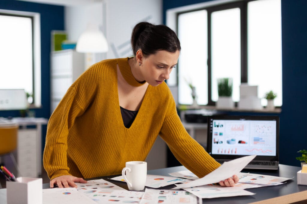 Das Foto zeigt eine Geschäftsfrau in einem Büro, die in ein Papierdokument vertieft ist. Sie trägt einen senfgelben Pullover und ihr Blick ist auf das Papier in ihren Händen gerichtet. Die Büroeinrichtung ist modern und hell. Auf ihrem Schreibtisch steht ein Laptop mit einer bunten Reihe von Diagrammen und Grafiken, die darauf hindeuten, dass sie Daten analysiert oder eine spezielle Software wie den Facebook Business Manager benutzt. Neben ihr steht eine weiße Tasse und auf dem Schreibtisch liegen weitere Dokumente und Diagramme, die auf einen sorgfältigen Arbeitsprozess schließen lassen. Die aufmerksame Körperhaltung der Frau und die Analysetools auf dem Bildschirm ihres Laptops lassen darauf schließen, dass sie sich intensiv mit Unternehmensanalysen oder strategischer Planung beschäftigt.