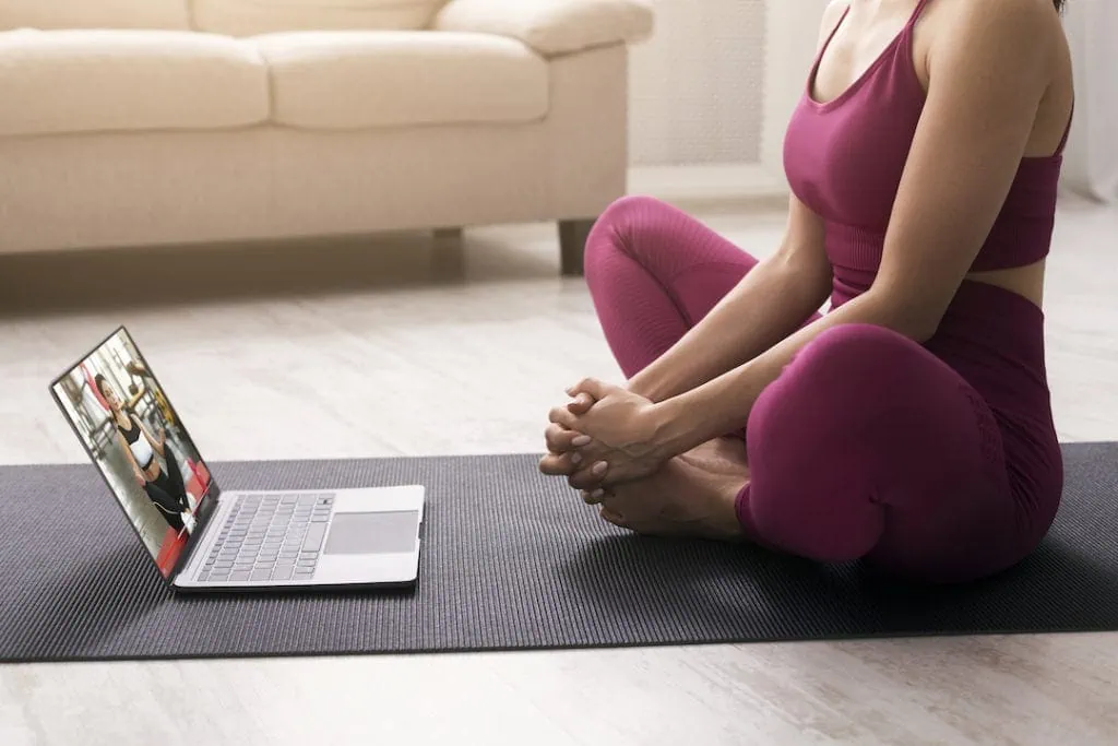 Social Media Kanäle: Eine junge Frau meditiert zu Hause im Lotussitz, während sie einen Online-Yogakurs auf Youtube besucht.