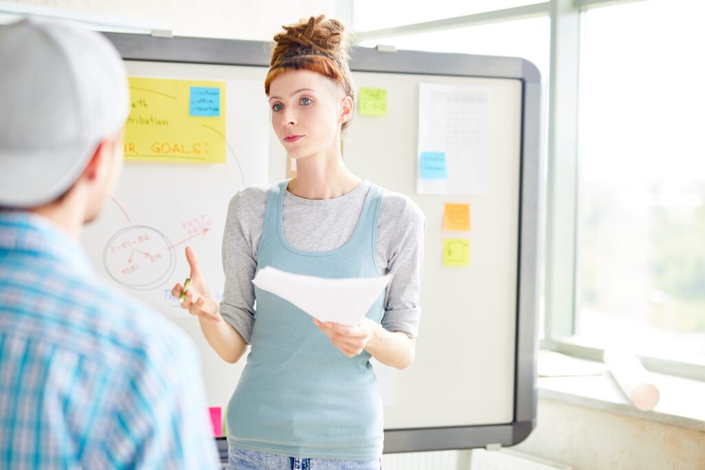 Seriöse, selbstbewusste Vermarkterin mit kurzem Pony, die mit der Hand gestikuliert und einen Kollegen anschaut, während sie eine Content Marketing Strategie zur Umsatzsteigerung diskutiert