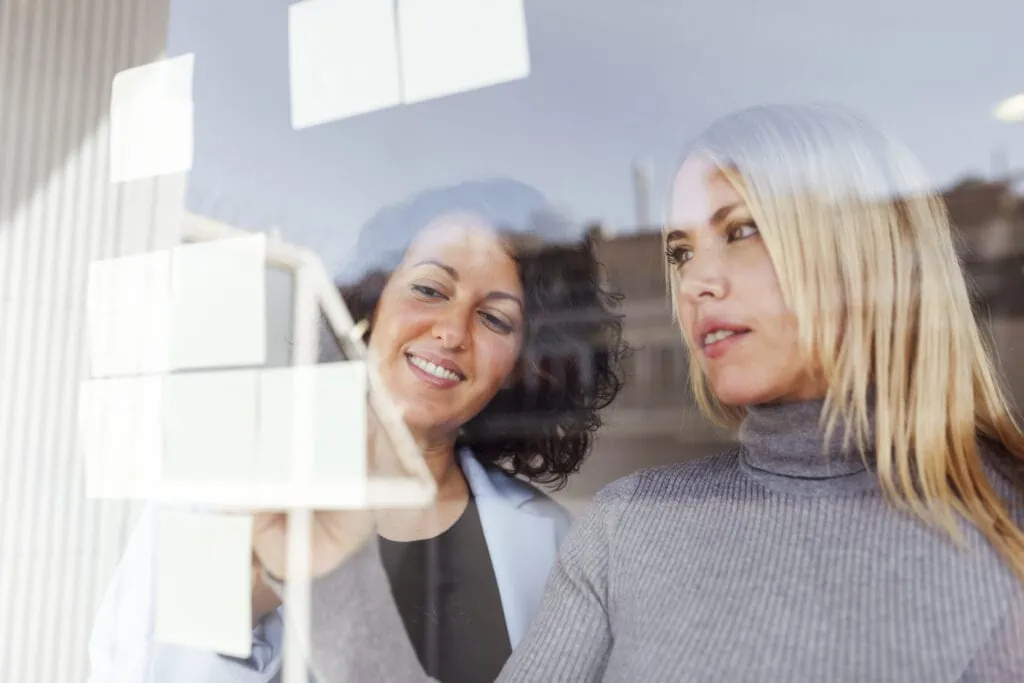 Marketing Beratung für Unternehmen: Geschäftsfrauen, die einen Plan erstellen, kleben Klebeetiketten an ein Fenster im Coworking Space.