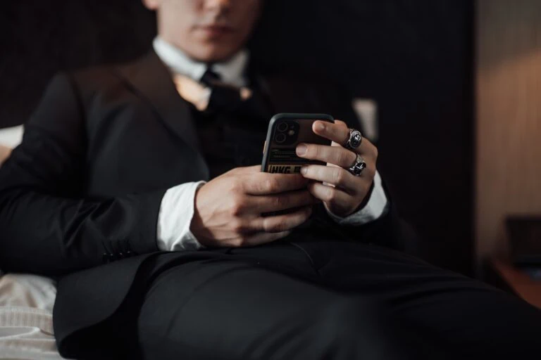 TikTok: Das Foto zeigt eine Person in einem formellen schwarzen Anzug mit einer weißen Hemdbluse und einer schwarz-weißen Krawatte. Die Person hält ein Smartphone in den Händen und scheint darauf zu tippen oder zu lesen. Man kann den oberen Teil des Körpers und die Hände sehen, aber das Gesicht ist nicht sichtbar, da der Kopf nicht im Bild ist. Die Person trägt mehrere Ringe an den Fingern, darunter einen auffälligen Ring mit einem großen Emblem am Zeigefinger. Der Hintergrund ist unscharf, sodass der Fokus auf den Händen und dem Smartphone liegt. Die Szene wirkt durch die Kleidung und die Accessoires stilvoll und modern.