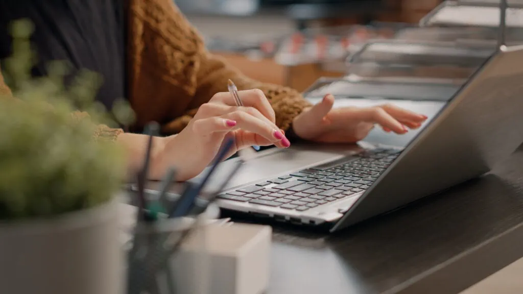 Das Foto zeigt eine Nahaufnahme der Hände einer Geschäftsfrau, die in einer Marketingagentur an einem Laptop arbeitet und Daten analysiert, um die Customer Journey zu optimieren. Eine Hand tippt auf der Tastatur des Laptops, während die andere einen Stift hält, was darauf hindeutet, dass sie abwechselnd digitale Analysen und Notizen macht. Ihre Fingernägel sind sorgfältig manikürt und rosa lackiert, und sie trägt eine braune Strickjacke, die ihrer professionellen Erscheinung einen Hauch von Wärme und Stil verleiht. Der Laptop steht auf einem schlanken, modernen Schreibtisch, daneben eine Tasse mit verschiedenen Stiften, was auf einen organisierten und effizienten Arbeitsplatz hindeutet. Im Vordergrund ist ein Hauch von Grün zu sehen, vielleicht eine Büropflanze, die der Umgebung einen Hauch von Leben und Lebendigkeit verleiht. Die Konzentration auf die Hände und die Arbeitswerkzeuge anstelle des Gesichts betont die Handlung und die zu erledigende Aufgabe und unterstreicht die detaillierte und konzentrierte Arbeit, die in die Analyse und Optimierung des Kundenerlebnisses einfließt.