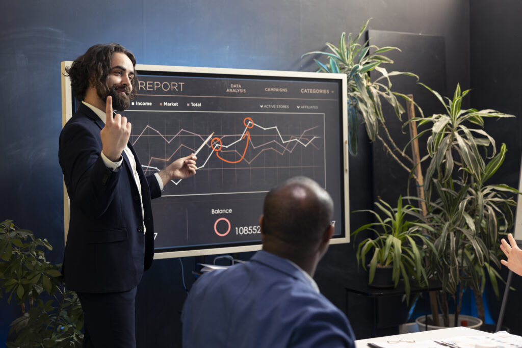 Ein Geschäftsmann in einem Anzug präsentiert eine Marketinganalyse auf einem großen Bildschirm mit Finanz- und Kampagnendiagrammen. Er erklärt eine Strategie, während Kollegen ihm aufmerksam zuhören.