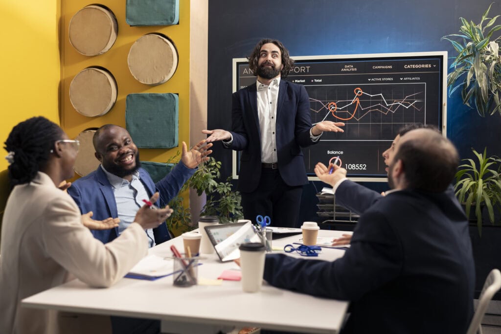 Ein Geschäftsmann in einem Anzug steht vor einem großen Bildschirm mit Marketing-Daten und erklärt eine Strategie mit einer humorvollen Geste. Das Team lacht und diskutiert lebhaft am Konferenztisch.