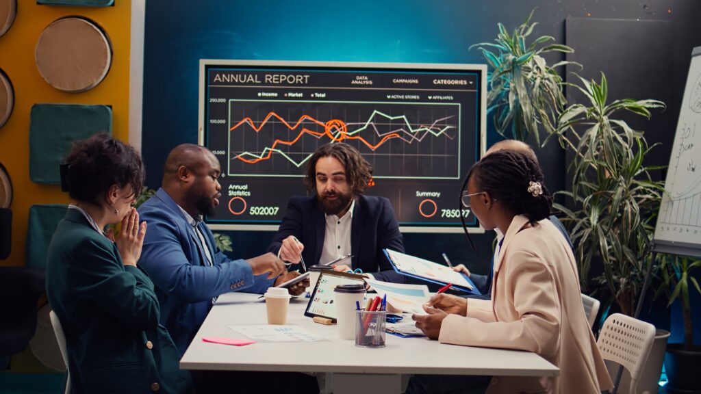 Ein Team von Geschäftsleuten sitzt an einem Konferenztisch und analysiert Marketingdaten. Im Hintergrund zeigt ein großer Bildschirm ein Annual Report-Diagramm mit Finanz- und Marktstatistiken, während der Teamleiter eine Strategie präsentiert.