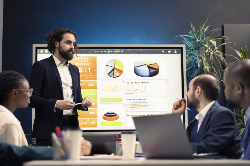 Ein Geschäftsmann in einem dunklen Anzug präsentiert eine Business-Analytics-Strategie vor einem großen Bildschirm mit Diagrammen, Umsatzanalysen und Markttrends. Das Team am Konferenztisch hört aufmerksam zu und diskutiert die Daten.
