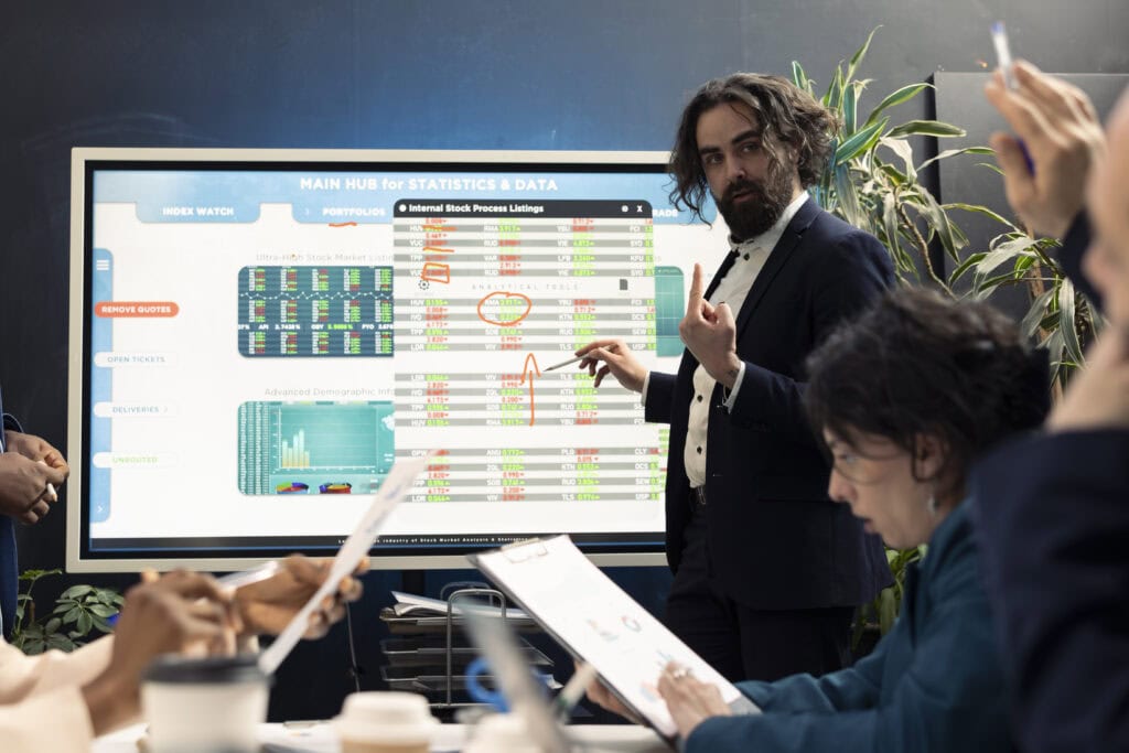 Ein Geschäftsmann in einem dunklen Anzug präsentiert eine Analyse von Finanz- und Marktstatistiken vor einem großen Bildschirm mit hervorgehobenen Datenpunkten. Das Team am Konferenztisch verfolgt die Präsentation und analysiert Dokumente mit Diagrammen.
