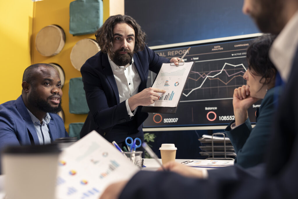 Ein Geschäftsmann in einem Anzug hält eine Präsentation über Marketing-Analytics-Daten, zeigt auf ein Diagramm und erklärt eine Strategie. Im Hintergrund ein großer Bildschirm mit Finanz- und Kampagnendaten, während Kollegen aufmerksam zuhören.