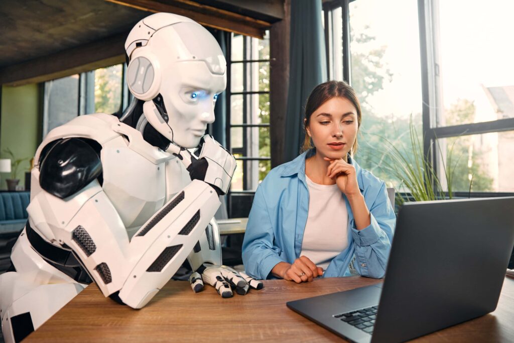KI-Agenten: Eine junge Frau und ein humanoider Roboter arbeiten gemeinsam an einem Laptop in einem modernen Büro. Eine Zusammenarbeit zwischen Mensch und künstlicher Intelligenz.