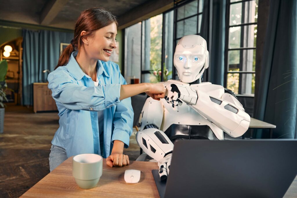 KI-Agenten im Unternehmen: Eine junge Frau arbeitet mit einem humanoiden Roboter an einem Laptop in einem modernen Büro und hält dabei eine Faust an die Faust. Dies symbolisiert die Zusammenarbeit zwischen Menschen und künstlicher Intelligenz.