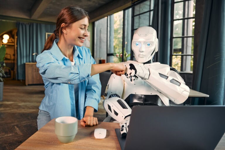 KI-Agenten im Unternehmen: Eine junge Frau arbeitet mit einem humanoiden Roboter an einem Laptop in einem modernen Büro und hält dabei eine Faust an die Faust. Dies symbolisiert die Zusammenarbeit zwischen Menschen und künstlicher Intelligenz.